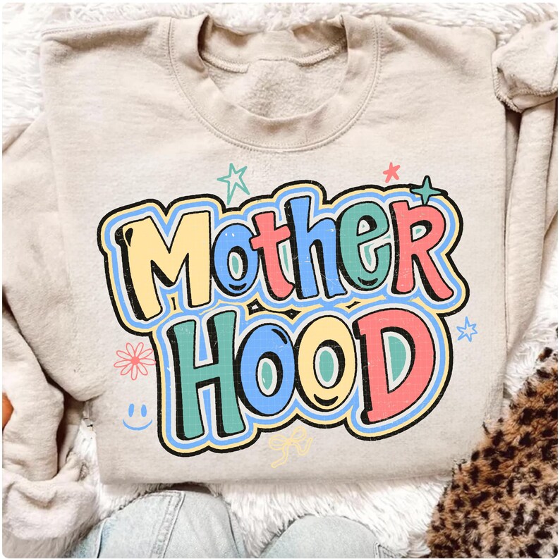 May include: Beige sweatshirt featuring the words "Mother Hood" in a colourful, retro font. The text is outlined in black and decorated with stars, a flower, and a smiley face. The sweatshirt has a crew neck and long sleeves.