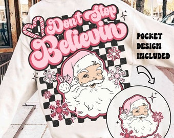 May include: Cream-colored sweatshirt featuring a retro design with the words "Don't Stop Believin" in pink. The design includes a winking Santa Claus, snowflakes, and a checkerboard pattern. The pocket design is also included.