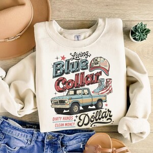 May include: Cream-colored sweatshirt with a graphic design featuring a blue and gold pickup truck, cowboy boots, and the text "Living Blue Collar Dollar." The design includes the phrases "Dirty Hands, Clean Money."