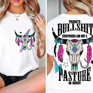 May include: White t-shirt with a graphic of a cow skull with turquoise and pink feathers and the text "There's Bullshit Everywhere and Not a Pasture in Sight".