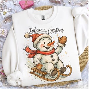 Op de afbeelding: Witte sweatshirt met een sneeuwpopontwerp. De sneeuwpop draagt een rode hoed, sjaal en wanten en zit op een houten slee. De tekst "Believe in the magic of Christmas" staat erboven. Een feestelijk kerstkledingstuk.