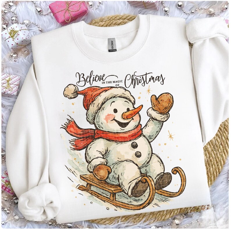 Op de afbeelding: Witte sweatshirt met een sneeuwpopontwerp. De sneeuwpop draagt een rode hoed, sjaal en wanten en zit op een houten slee. De tekst "Believe in the magic of Christmas" staat erboven. Een feestelijk kerstkledingstuk.