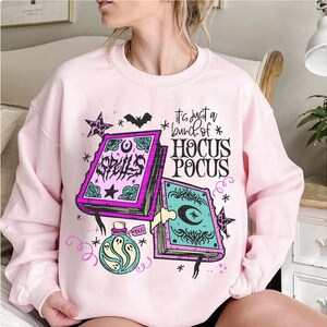 May include: A pale pink sweatshirt with a graphic design. The design features the text "It's just a bunch of Hocus Pocus", two spell books, stars, a bat, and a potion bottle with a ghost. The books are purple and teal.