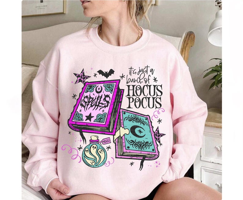 May include: A pale pink sweatshirt with a graphic design. The design features the text "It's just a bunch of Hocus Pocus", two spell books, stars, a bat, and a potion bottle with a ghost. The books are purple and teal.