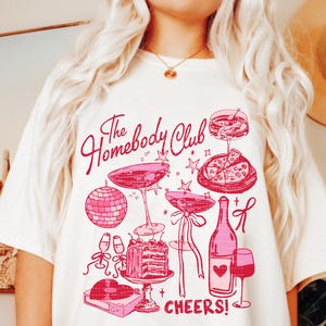 May include: White t-shirt with a pink graphic design that says "The Homebody Club" and features illustrations of a disco ball, champagne flutes, a bottle of wine, a glass of wine, a slice of pizza, and a cake. The text "CHEERS!" is at the bottom of the design.