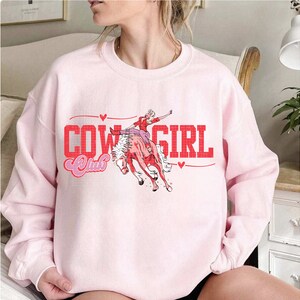 May include: Pink sweatshirt featuring a graphic of a woman riding a horse and the text "Cowgirl Club" in red.