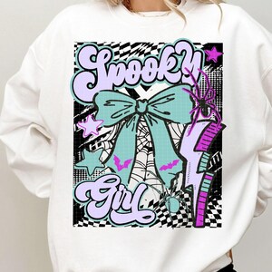 May include: White sweatshirt with a colorful graphic that says "Spooky Girl." The design includes a large teal bow, bats, stars, a spider, and a lightning bolt. The background has a black and white checkered pattern.