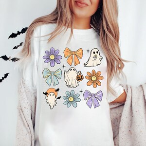May include: White t-shirt with a colorful Halloween design featuring ghosts, bows, and flowers. The design includes a ghost holding a pumpkin, a ghost with a flower, and a ghost with bats.