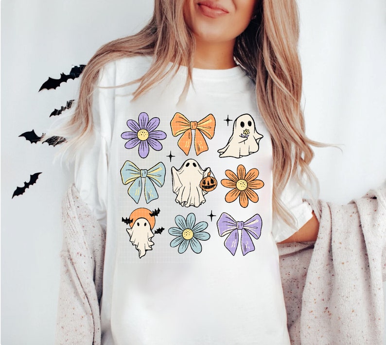 May include: White t-shirt with a colorful Halloween design featuring ghosts, bows, and flowers. The design includes a ghost holding a pumpkin, a ghost with a flower, and a ghost with bats.
