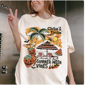 May include: Cream-colored t-shirt with a colorful graphic featuring a palm tree, sun, beach umbrella, and watermelon slice. The text reads "Chillin' & Thrillin'" and "Summer-ween Vibes". The design includes bats and jack-o'-lantern strawberries.
