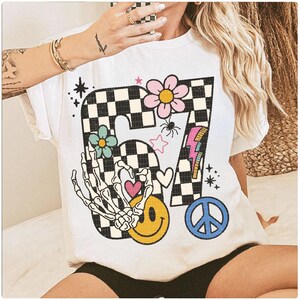 May include: White t-shirt featuring a colorful graphic design. The design includes a checkered number 67, flowers, a skeleton hand, a smiley face, a peace sign, and a lightning bolt. The shirt has short sleeves and a crew neck.
