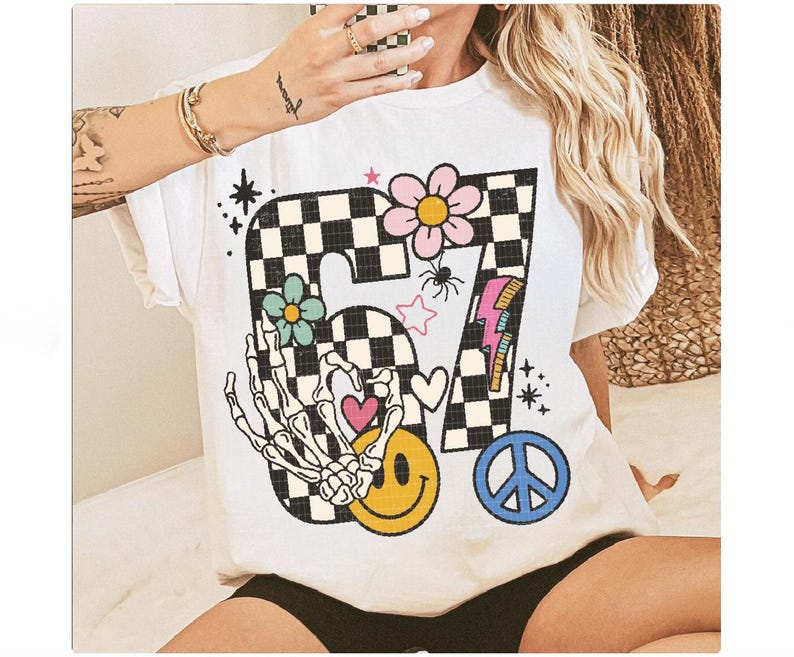 May include: White t-shirt featuring a colorful graphic design. The design includes a checkered number 67, flowers, a skeleton hand, a smiley face, a peace sign, and a lightning bolt. The shirt has short sleeves and a crew neck.