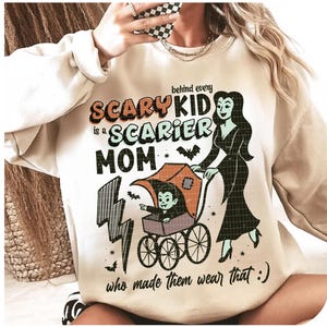 May include: Beige sweatshirt with a Halloween-themed graphic. The design features the text "Behind every Scary Kid is a Scarier Mom" with a cartoon illustration of a vampire child in a stroller and a woman. Bats and lightning bolts are also included.