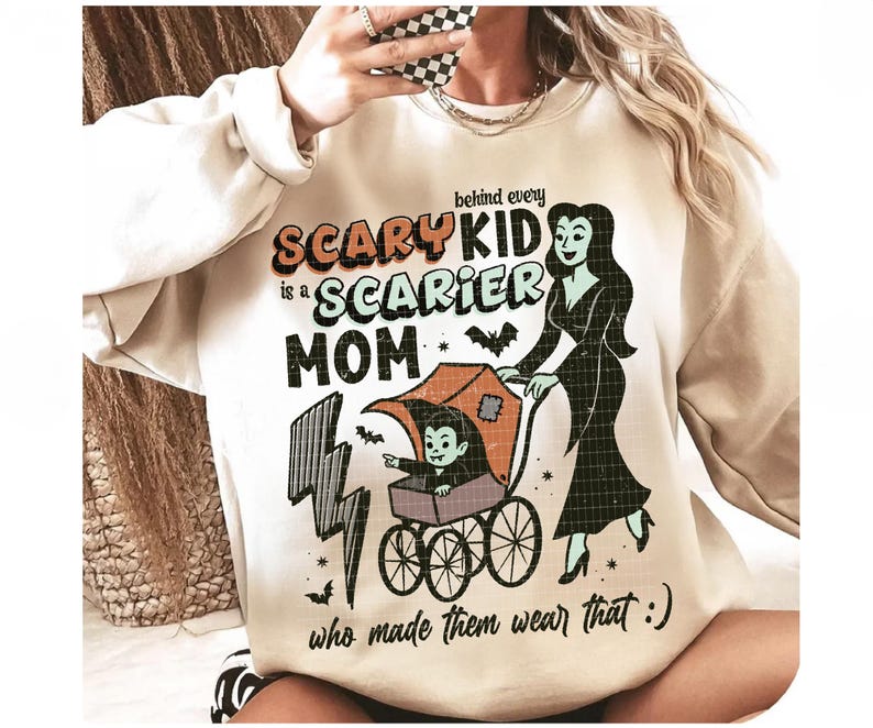 May include: Beige sweatshirt with a Halloween-themed graphic. The design features the text "Behind every Scary Kid is a Scarier Mom" with a cartoon illustration of a vampire child in a stroller and a woman. Bats and lightning bolts are also included.
