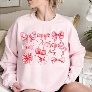 May include: Pink sweatshirt with a graphic design of red bows, cherries, and stars.
