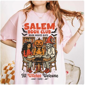 Może przedstawiać: Jasnor&oacute;żowa koszulka z napisem "Salem Book Club" i grafiką przedstawiającą trzy postacie wok&oacute;ł stołu. Postacie to wiedźma, dynia i szkielet. Tekst "All Witches Welcome - 1692" jest r&oacute;wnież zawarty.
