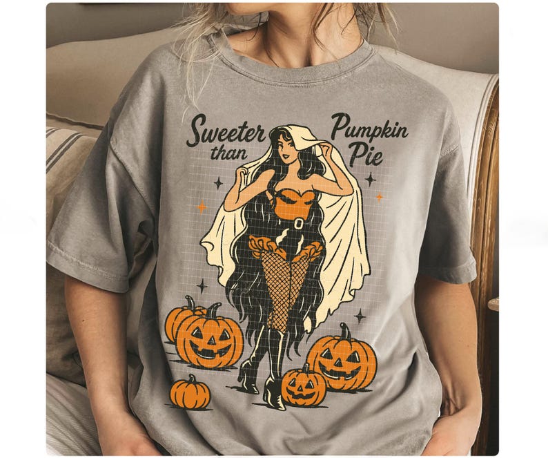 Peut inclure: T-shirt gris avec un motif sur le th&egrave;me d'Halloween. Le design pr&eacute;sente une femme de style pin-up en costume, entour&eacute;e de citrouilles. Le texte "Sweeter than Pumpkin Pie" est au-dessus de l'illustration. Le t-shirt est de coupe ample.
