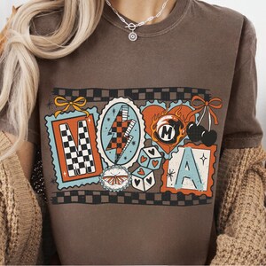 Op de afbeelding: Bruin t-shirt met een kleurrijk grafisch ontwerp. Het ontwerp bevat de letters "M" en "A" in postzegelachtige kaders, samen met andere retro-illustraties zoals een bliksemschicht en dobbelstenen.