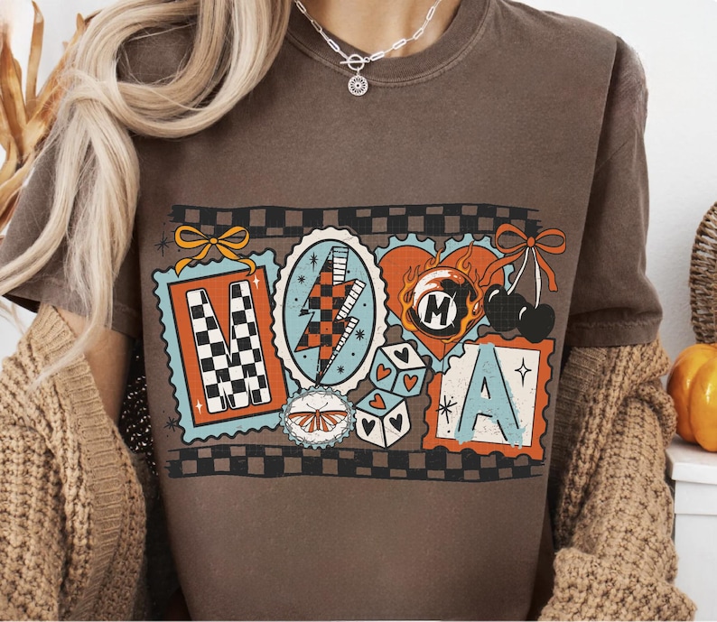 Op de afbeelding: Bruin t-shirt met een kleurrijk grafisch ontwerp. Het ontwerp bevat de letters "M" en "A" in postzegelachtige kaders, samen met andere retro-illustraties zoals een bliksemschicht en dobbelstenen.