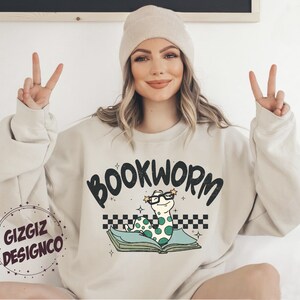Pu&ograve; includere: Una felpa beige chiaro con il testo "BOOKWORM" in lettere nere. Un verme dei cartoni animati con gli occhiali &egrave; seduto su un libro con un motivo a scacchi.