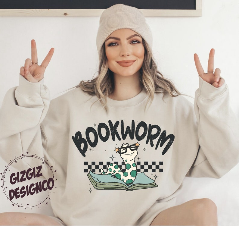 Pu&ograve; includere: Una felpa beige chiaro con il testo "BOOKWORM" in lettere nere. Un verme dei cartoni animati con gli occhiali &egrave; seduto su un libro con un motivo a scacchi.