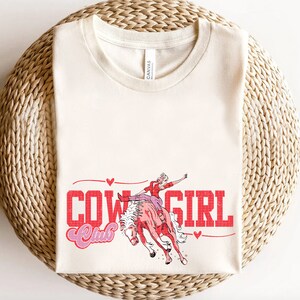 May include: A cream-coloured t-shirt with a red and pink graphic design that says "Cowgirl Club". The graphic features a woman riding a bucking horse.