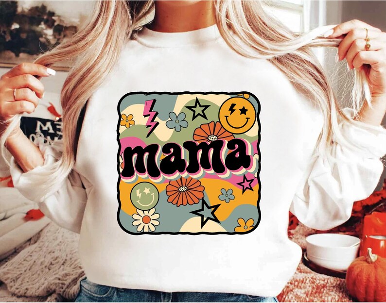 Op de afbeelding: Witte trui met een retro-grafisch ontwerp met het woord "mama" in een kleurrijk, golvend lettertype. Het ontwerp bevat bloemen, sterren en een smiley.