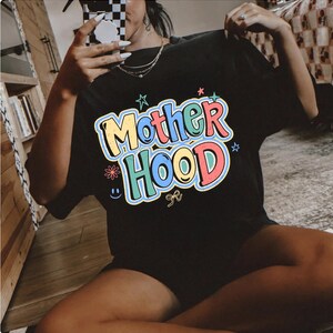 May include: Black t-shirt featuring the word "Motherhood" in a colourful, bubble-style font. The design includes stars, a flower, a smiley face, and a bow. The shirt is a casual, comfortable style.