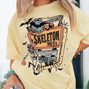 Puede incluir: Camiseta amarilla clara con un dise&ntilde;o retro de "The Skeleton Motel". El dise&ntilde;o incluye un esqueleto, murci&eacute;lagos y un letrero de motel con el texto "Old Graveyard Road" y "No Escape Since 1690".
