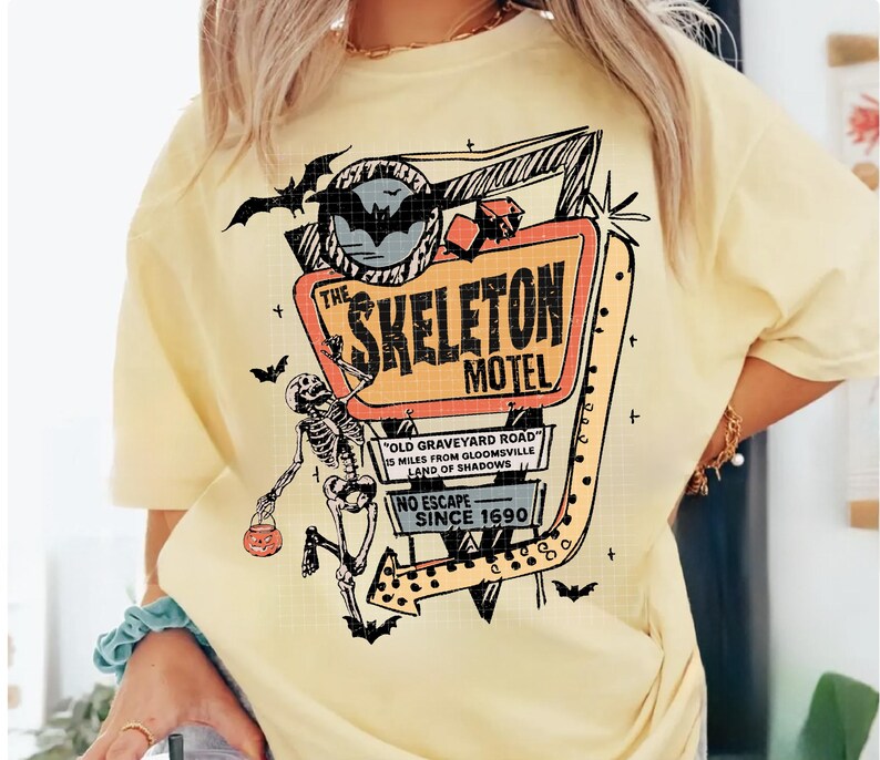 Puede incluir: Camiseta amarilla clara con un dise&ntilde;o retro de "The Skeleton Motel". El dise&ntilde;o incluye un esqueleto, murci&eacute;lagos y un letrero de motel con el texto "Old Graveyard Road" y "No Escape Since 1690".