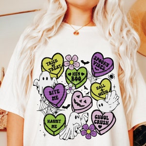May include: A white t-shirt with a colorful graphic design of candy hearts with Halloween-themed phrases like "Trick or Treat", "Bite Me", "Spook Vibes", and "Ghoul Crush".