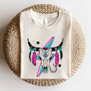 May include: A white t-shirt with a graphic of a turquoise and pink lightning bolt behind a black and white cow skull with turquoise and pink feathers and turquoise and pink floral accents.