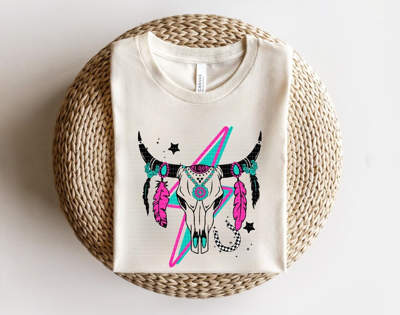 May include: A white t-shirt with a graphic of a turquoise and pink lightning bolt behind a black and white cow skull with turquoise and pink feathers and turquoise and pink floral accents.