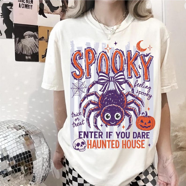 May include: A cream-colored t-shirt featuring a graphic print with the text SPOOKY, feeling spooky, trick or treat, and ENTER IF YOU DARE HAUNTED HOUSE. The design includes a purple spider with a bow, a spiderweb, a crescent moon, and a pumpkin.