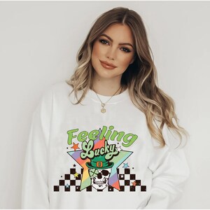 K&ouml;nnte beinhalten: Wei&szlig;er Pullover mit einem Regenbogen-Stern-Motiv und dem Text "Feeling Lucky" in Gr&uuml;n. Das Stern-Motiv zeigt einen Sch&auml;del, der einen gr&uuml;nen Koboldhut und eine Sonnenbrille tr&auml;gt.