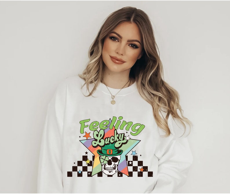 K&ouml;nnte beinhalten: Wei&szlig;er Pullover mit einem Regenbogen-Stern-Motiv und dem Text "Feeling Lucky" in Gr&uuml;n. Das Stern-Motiv zeigt einen Sch&auml;del, der einen gr&uuml;nen Koboldhut und eine Sonnenbrille tr&auml;gt.