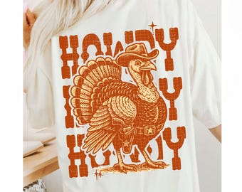 May include: Cream-colored t-shirt featuring a turkey wearing a cowboy hat and holster, with the word "HOWDY" in orange block letters. The turkey illustration is detailed with feathers and a belt. A fun, festive design.