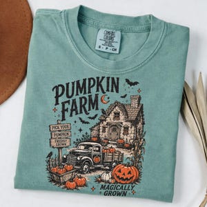 Peut inclure: Un t-shirt vert sauge avec une impression graphique repr&eacute;sentant une ferme de citrouilles, une maison, un vieux camion et des citrouilles. Le texte indique PUMPKIN FARM, PICK YOUR PUMPKIN FRESHLY GROWN et MAGICALLY GROWN. &Eacute;tiquette Comfort Colors.