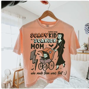May include: Peach-colored t-shirt with a Halloween-themed graphic. The design features the text "Scary Kid is a Scarier Mom" with a cartoon illustration of a vampire child in a stroller and a woman in a black dress.