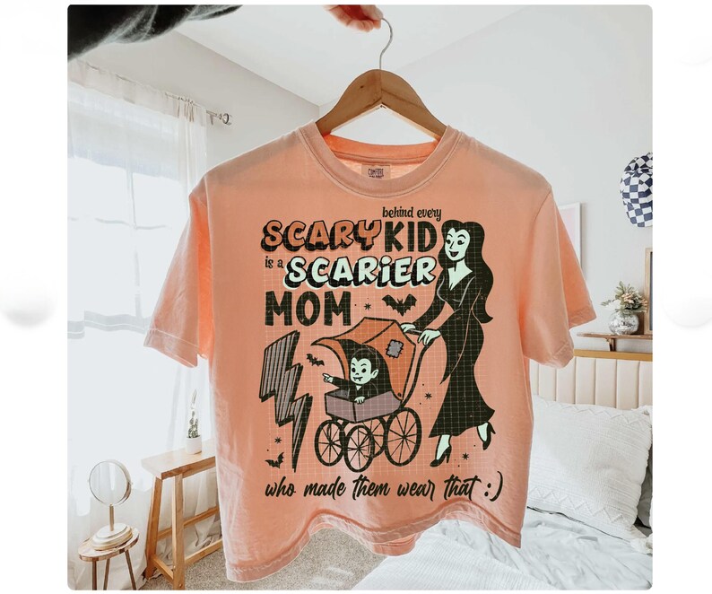 May include: Peach-colored t-shirt with a Halloween-themed graphic. The design features the text "Scary Kid is a Scarier Mom" with a cartoon illustration of a vampire child in a stroller and a woman in a black dress.
