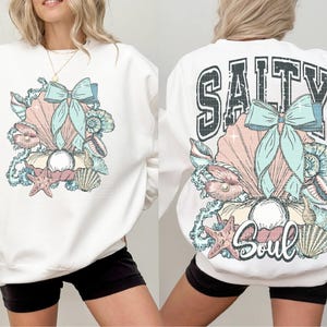Puede incluir: Sudadera blanca con un dise&ntilde;o de concha y lazo en la parte delantera y el texto "Salty Soul" con un dise&ntilde;o de concha y lazo en la parte trasera.