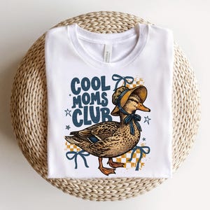 May include: White t-shirt featuring a duck wearing a bonnet and a blue bow. The text "COOL MOMS CLUB" is printed in blue with star accents. The duck is set against a yellow and white checkered background.
