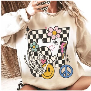 May include: Beige sweatshirt with a large graphic featuring a checkered "7" design, flowers, a skeleton hand, a smiley face, a peace sign, and a lightning bolt. The design incorporates pink, blue, yellow, and black elements, creating a retro, whimsical aesthetic.