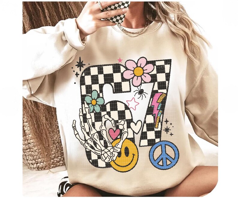 May include: Beige sweatshirt with a large graphic featuring a checkered "7" design, flowers, a skeleton hand, a smiley face, a peace sign, and a lightning bolt. The design incorporates pink, blue, yellow, and black elements, creating a retro, whimsical aesthetic.