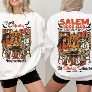 Może przedstawiać: Biała bluza z grafiką o tematyce Halloween. Prz&oacute;d przedstawia kresk&oacute;wkę trzech postaci wok&oacute;ł stołu z napisem "Meets Monthly Massachusetts". Tył m&oacute;wi "Salem Book Club" i "All Witches Welcome".