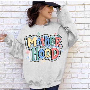 May include: A light grey sweatshirt featuring the word "Motherhood" in colourful bubble letters. The design includes stars, flowers, and a smiley face. The sweatshirt has long sleeves.