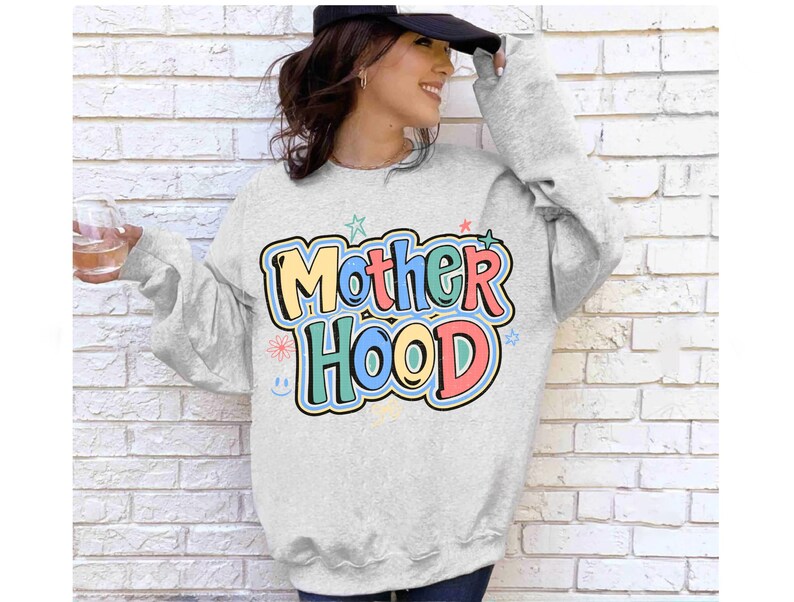 May include: A light grey sweatshirt featuring the word "Motherhood" in colourful bubble letters. The design includes stars, flowers, and a smiley face. The sweatshirt has long sleeves.