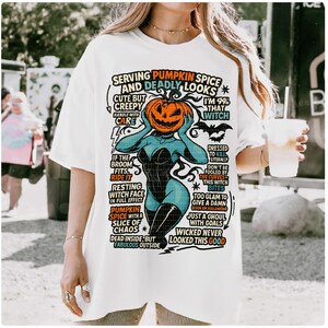 May include: White t-shirt with a Halloween-themed graphic. The design features a pumpkin-headed witch with text that reads "Serving Pumpkin Spice and Deadly Looks." The shirt has a relaxed fit and short sleeves.