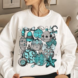 Peut inclure: Sweat-shirt blanc avec un motif d'ornements de No&euml;l repr&eacute;sentant des ornements bleus et gris avec des accents de cannes en sucre et d'&eacute;toiles.