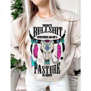 May include: A beige sweatshirt with a black and white graphic of a cow skull with turquoise and pink accents. The text "There's Bullshit Everywhere and Not a Pasture in Sight" is printed on the sweatshirt.